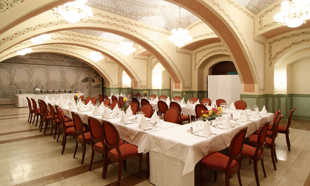 In U-Form aufgebaute Tische für eine Pensionsfeier im Lanner-Lehár-Saal des Wiener Rathauskellers. Mit weißen Tischtüchern und Servietten gedeckt, dazwischen Blumengestecke. Im hinteren Teil des Raums ein aufgebautes Buffet