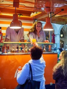Bar-Mitarbeiterin in einem Aperol-Schankwagen, die einem Gast ein Glas Aperol Spritz überreicht